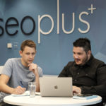 Two seoplus employees working and looking at a computer screen. Behind them is the seoplus+ logo on the wall.