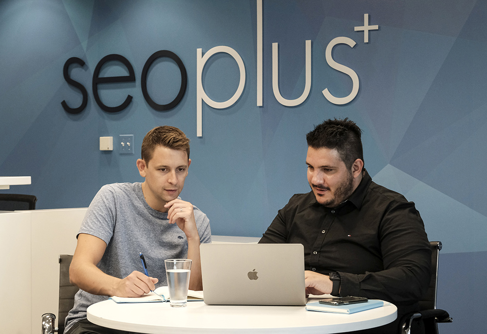 Two seoplus employees working and looking at a computer screen. Behind them is the seoplus+ logo on the wall.